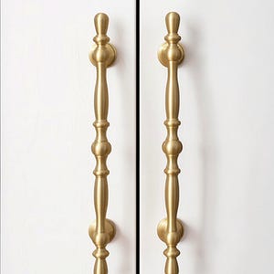 May include: Two brass-colored cabinet pulls with a decorative design. Each pull has a long, vertical handle with a series of rounded and cylindrical elements. The pulls are mounted on a white surface. The text "CraftedHooksDecor" is in the upper right corner.