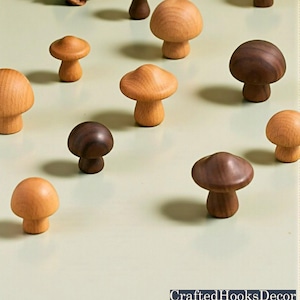 以下が含まれることがあります： さまざまな茶色の木製キノコ型装飾ノブのコレクション。ノブは明るい緑色の表面に配置されています。「CraftedHooksDecor」の文字が右下に表示されています。