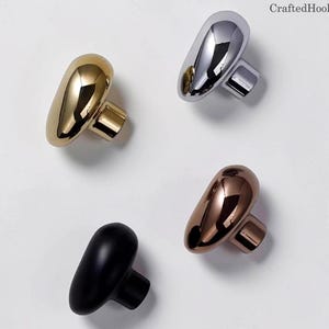 May include: Four modern cabinet knobs in gold, silver, bronze, and black. Each knob has a smooth, rounded shape with a rectangular base. The knobs are displayed against a white background, showcasing their reflective surfaces.