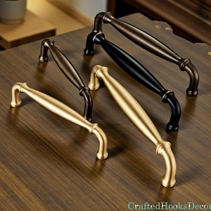 Puede incluir: Una variedad de tiradores de armario en acabados dorado, negro y bronce. Los tiradores tienen un diseño curvo y están colocados sobre una superficie de madera. El texto "CraftedHooksDecor" es visible en la esquina inferior derecha.