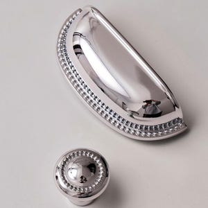 May include: A polished silver cabinet pull and matching knob. The pull is a half-moon shape with a beaded border. The knob is round with a similar beaded design. Both pieces reflect the surrounding environment.