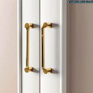 Op de afbeelding: Twee goudkleurige kastgrepen zijn gemonteerd op een witte kastdeur. De handgrepen zijn lang en slank, met een gebogen ontwerp aan de bovenkant. De tekst "CraftedHooksDecor" staat in de rechterbovenhoek.