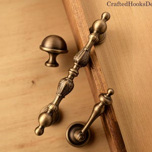 Pode incluir: Uma variedade de ferragens de armário em bronze antigo, incluindo um botão, uma puxador e um puxador menor. O hardware é exibido em uma superfície de madeira. O texto "CraftedHooksDecor" é visível no canto superior direito.