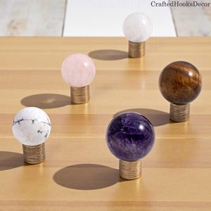 May include: Five decorative cabinet knobs with spherical tops. The knobs feature a variety of materials, including white marble, rose quartz, amethyst, clear quartz, and wood. Each knob is mounted on a short, cylindrical brass base. The CraftedHooksDecor logo is in the top right corner.