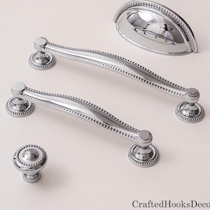 May include: A collection of polished chrome cabinet hardware, including two long pulls, a cup pull, and a round knob. The hardware features a beaded edge design and a reflective surface, ideal for kitchen or bathroom renovations.