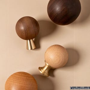 May include: A collection of wooden cabinet knobs with brass bases. The knobs are various shades of brown, from light to dark, and are spherical in shape. The brass bases have a conical design. The knobs are arranged on a neutral-toned surface.