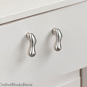 Może przedstawiać: Zbliżenie dwóch srebrnych uchwytów do szafek o unikalnym, organicznym kształcie. Uchwyty są zamontowane na białej szufladzie szafki, oferując nowoczesny i stylowy element designu. Uchwyty wykonane są z błyszczącego, odblaskowego metalu.