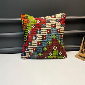 May include: A square, patterned pillow with a vibrant mix of colors. The pillow features a woven design with red, blue, green, and white geometric shapes. The pillow is placed on a white surface with a dark gray background.