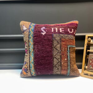 May include: Square decorative pillow with a patchwork design. The pillow features a deep burgundy center with colorful geometric patterns in shades of brown, teal, orange, and purple. The word "NEV" is visible on the top of the pillow.