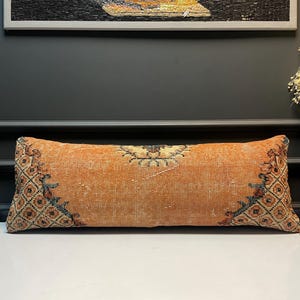 Può includere: Cuscino lombare rettangolare arancione con un aspetto sbiadito e usurato. Il cuscino presenta un design geometrico centrale e estremità decorate con motivi arancioni, blu e neri. Progettato per uso decorativo.