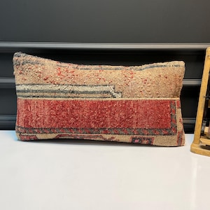 Op de afbeelding: Rechthoekig kussen in vintage-stijl met een patchwork-ontwerp. Het bovenste gedeelte is beige met rode accenten, het midden heeft grijze strepen en de onderkant is effen rood. Het kussen is gemaakt van een textiel.