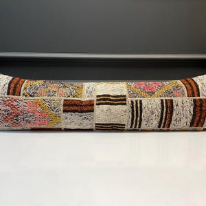 Könnte beinhalten: Ein langes, rechteckiges Kissen mit einem Patchwork-Design. Das Kissen weist eine Vielzahl von Farben auf, darunter Beige, Braun, Rosa und Gelb. Das Kissen wird wahrscheinlich zu Dekorationszwecken auf einem Bett oder Sofa verwendet.