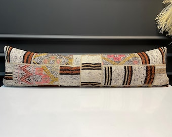 May include: A long, rectangular cushion with a patchwork design. The cushion features a variety of colours, including beige, brown, pink, and yellow. The cushion is likely used for decorative purposes on a bed or sofa.