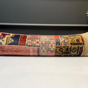 May include: A long, rectangular cushion with a patchwork design. The cushion features a variety of colours, including red, blue, pink, and beige. The patchwork design is made up of different patterns and textures, creating a unique and eye-catching look.