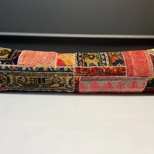 Puede incluir: Un largo cojín rectangular con un diseño de patchwork. El cojín presenta varios parches rectangulares de diferentes colores y patrones, incluyendo rojo, rosa, azul y dorado. El cojín se utiliza probablemente con fines decorativos.