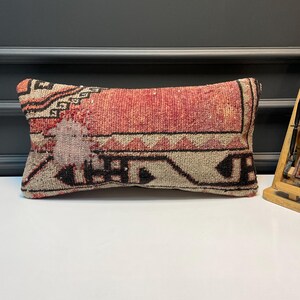 May include: Rectangular decorative pillow with a woven design. The pillow features a central section in shades of red and pink, bordered by a beige and black geometric pattern. The pillow is placed on a white surface.