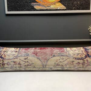 May include: A long, rectangular pillow with a vintage rug design. The pillow features a central maroon section with beige and blue accents. The overall design has a distressed, antique appearance, suggesting it is a decorative item.