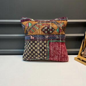 May include: A small, decorative pillow with a patchwork design. The pillow features a mix of colors including burgundy, gold, blue, and green. The design includes geometric patterns and floral motifs, creating a vintage or bohemian aesthetic.