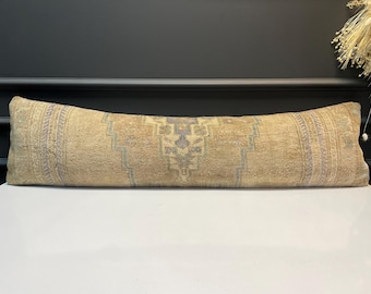 May include: A long, rectangular cushion with a beige and faded blue geometric pattern. The cushion has a vintage, distressed look, with a central design and border details. It rests on a white surface.