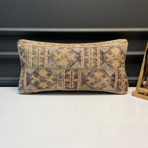 Pode incluir: Almofada decorativa retangular com um design tecido em tons de bege, laranja, verde e roxo. A almofada tem uma superfície texturizada e uma estética vintage, adequada para decoração de casa.