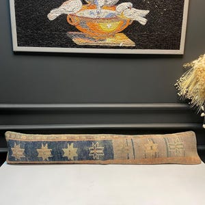 May include: A long, rectangular pillow with a woven design. The pillow features a blue and beige color scheme with geometric patterns. The pillow is placed on a white surface, with a mosaic artwork of birds in the background.
