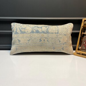May include: Rectangular beige pillow with a faded blue floral pattern. The pillow has a vintage, distressed look, with a soft, textured appearance. The design features a central medallion and border details, suggesting a rug-inspired aesthetic.