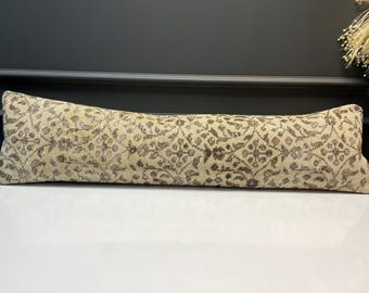 May include: A long, rectangular cushion with a beige background and a repeating floral pattern in shades of brown. The cushion is placed on a white surface, with a dark background.