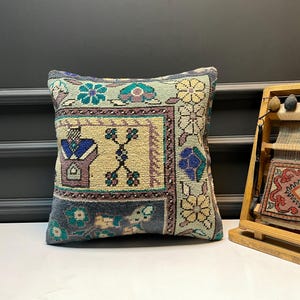 Puede incluir: Cojín decorativo cuadrado con un diseño vintage. El cojín presenta un panel central beige con un motivo en forma de cruz, rodeado de bordes florales y geométricos en tonos azules, verdes y morados. El cojín está colocado sobre una superficie blanca.
