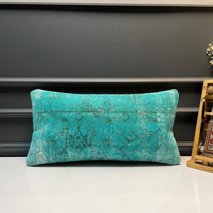 Puede incluir: Un cojín rectangular turquesa con un patrón descolorido y desgastado. El cojín tiene una apariencia vintage y antigua, con una mezcla de tonos turquesa claros y oscuros. El diseño presenta sutiles acentos más oscuros.