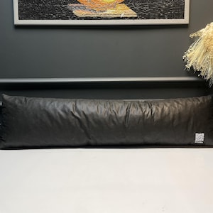May include: A long, black bolster cushion with a textured surface. The cushion is rectangular and appears to be filled with a soft material. The cushion is resting on a white surface.