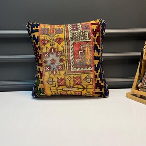 May include: Square decorative pillow with a vibrant, geometric pattern in yellow, red, and blue. The pillow features a detailed, woven design with a dark blue border. The pillow is placed on a white surface.