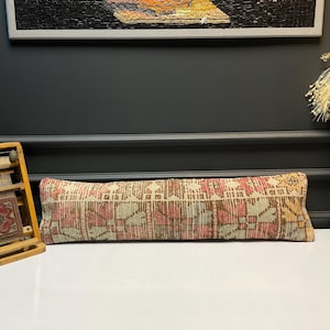 May include: A rectangular, patterned pillow with a beige background and red, brown, and green geometric designs. The pillow is long and narrow, likely for lumbar support or decorative use. The pillow is placed on a white surface.