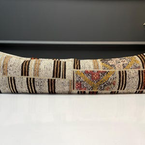 May include: A long, rectangular cushion with a patchwork design. It has a cream background with vertical brown stripes and geometric patterns in red, yellow, and brown. The cushion is placed on a white surface.