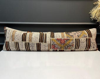 May include: A long, rectangular cushion with a patchwork design. It has a cream background with vertical brown stripes and geometric patterns in red, yellow, and brown. The cushion is placed on a white surface.