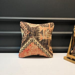 May include: A small, square decorative pillow with a vintage, patterned design. The pillow features a mix of colors, including brown, peach, and white, with geometric and floral motifs. The pillow is likely made of a woven fabric.