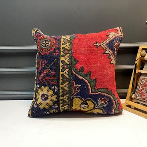 May include: Square decorative pillow with a vibrant, patterned design. The pillow features a red central field with a detailed border in navy blue, yellow, and green. The pillow's corners have a unique shape, adding to its decorative appeal.
