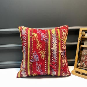 May include: A square, red decorative pillow with a woven design. The pillow features vertical stripes in yellow and white, with floral and geometric patterns in purple and green. The pillow is likely handmade.