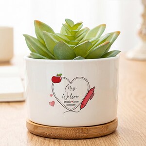 May include: A white ceramic cylindrical planter with a bamboo base containing a green succulent. The planter features a heart-shaped graphic with a red apple, a red pen, and the text "Mrs. Wilson" above the words "teach love inspire".