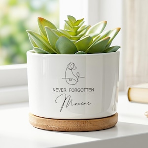 May include: A white ceramic cylindrical planter containing a green succulent. The front features a black line-art drawing of a dog, the text "NEVER FORGOTTEN", and the name "Maxine" in script. The planter sits on a circular bamboo base.