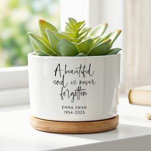 Op de afbeelding: Een witte cilindervormige keramische plantenpot met een bamboe onderzetter en een groene vetplant. De pot is voorzien van de zwarte tekst A beautiful soul is never forgotten, gevolgd door de naam EMMA SWAN en de jaartallen 1954-2025.