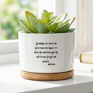 Peut inclure: Un pot de fleurs cylindrique en céramique blanche contenant une plante succulente verte. Le pot présente un texte imprimé en noir qui dit : Friendship isn't about who you've known the longest, it's about who walked into your life, said I'm here for you and proved it. Il repose sur une base ronde en bambou.
