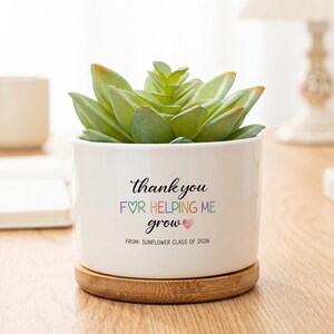 May include: A white ceramic planter pot containing a green succulent plant, resting on a circular bamboo wood base. The front of the pot features the black text "thank you FOR HELPING ME grow" with a small heart, and "FROM: SUNFLOWER CLASS OF 2026".