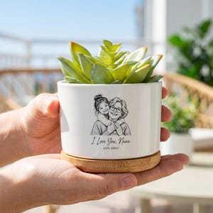 Puede incluir: Una maceta cilíndrica de cerámica blanca con base de bambú que contiene una suculenta verde. El frente presenta una ilustración de líneas negras de un niño abrazando a un adulto, con el texto "I Love You, Nana" y "LOVE, EMILY" impreso debajo.