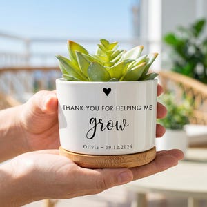 Könnte beinhalten: Ein weißer Keramikübertopf mit Bambussockel und einer grünen Sukkulente. Der Übertopf ist mit einem schwarzen Herzsymbol, dem Text "THANK YOU FOR HELPING ME grow" sowie dem personalisierten Text "Olivia • 09.12.2026" in schwarzer Schrift bedruckt.
