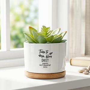 May include: A white ceramic cylindrical planter with a bamboo saucer base containing a green succulent. The planter features black printed text that reads Time to bloom DAISY HAPPY RETIREMENT 2026 with small leaf illustrations on the front surface.