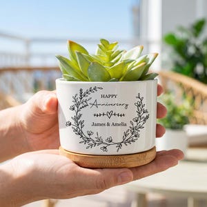May include: A white ceramic planter with a bamboo saucer containing a green succulent. The planter features a black printed floral wreath design with the text "Happy Anniversary" and the names "James & Amelia" centered inside the wreath.