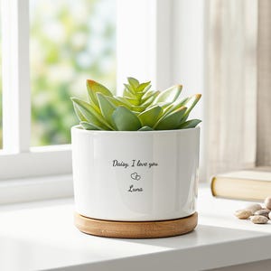 May include: A white cylindrical ceramic planter containing a green succulent plant. The planter features the black printed text "Daisy, I love you" above two small heart outlines and the name "Luna". It sits on a circular light brown wooden base.