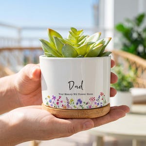 Może przedstawiać: Cylindryczna doniczka z białej ceramiki z bambusową podstawką zawierająca zielony sukulent. Doniczka posiada czarny napis Dad oraz Your Memory Will Forever Bloom nad kolorową obwódką z motywem drobnych polnych kwiatów.