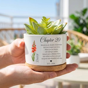 Puede incluir: Una maceta de cerámica blanca con base de bambú que contiene una suculenta verde. La maceta presenta un gráfico floral impreso, el texto "Chapter 20", un poema sobre recuerdos, el nombre "Daisy", un icono de corazón y "EST. 2006".