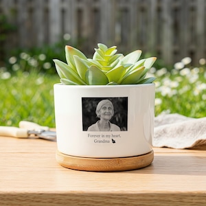 May include: A white ceramic planter with a bamboo base containing a green succulent. The front features a black and white photograph of an elderly person with the text "Forever in my heart, Grandma" and a small butterfly icon printed below it.